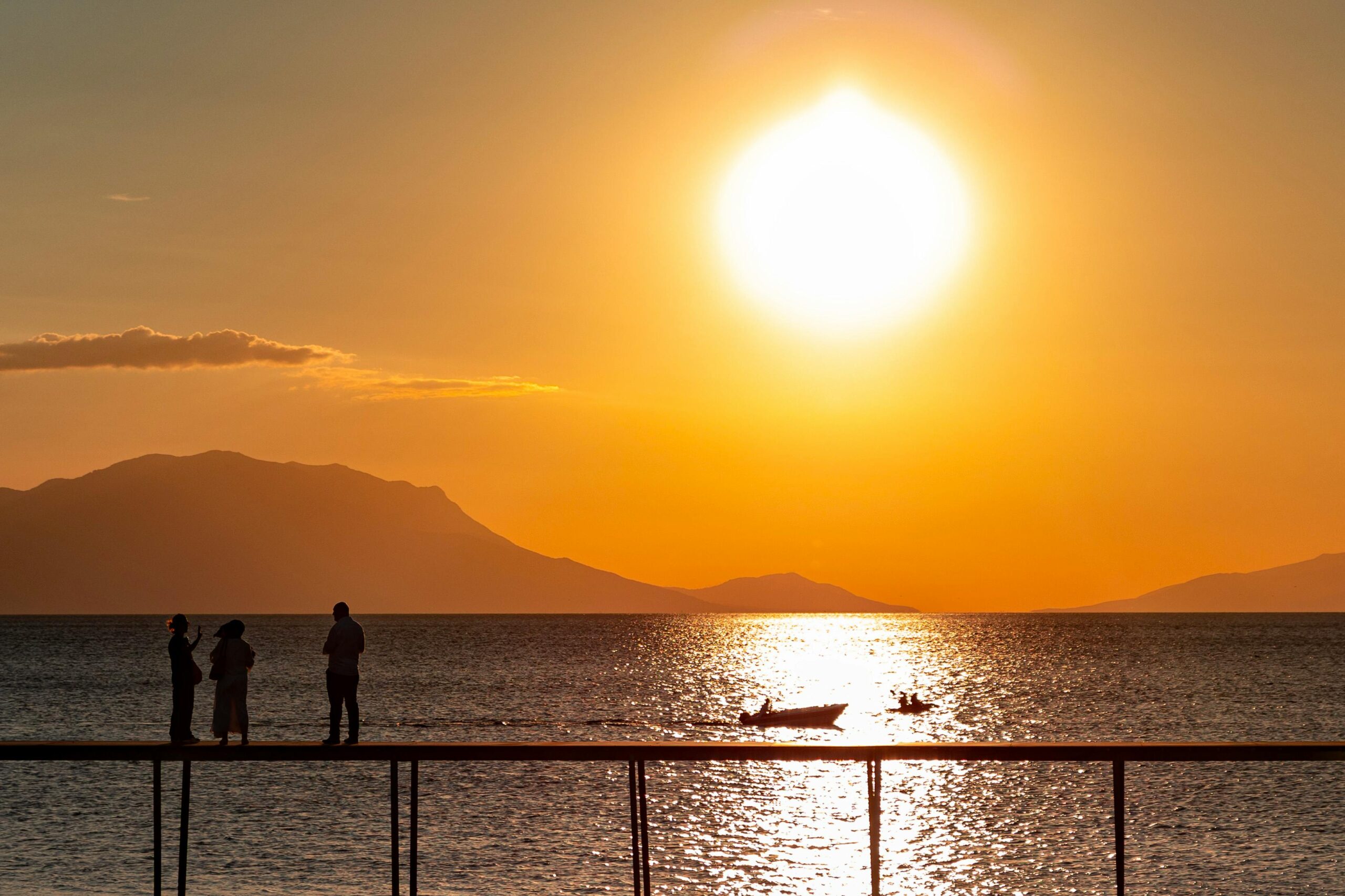 Silhouettes of people on a pier overlooking a shimmering sea during a vibrant golden sunset, with small boats on the water and layered mountains in the distance.
